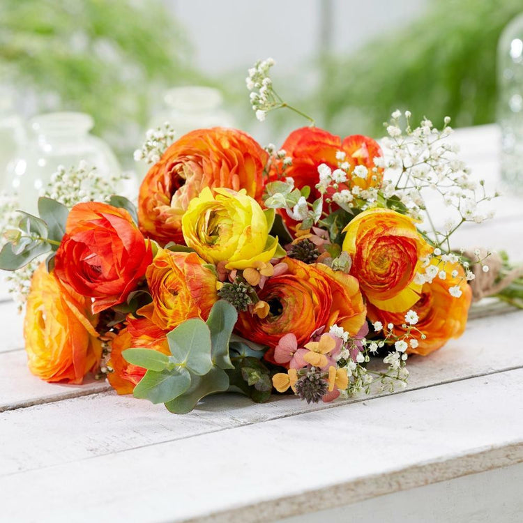 A bouquet of yellow, red, and orange Aviv ranunculus flowers, mixed with greens and baby's breath, lying on a white table.